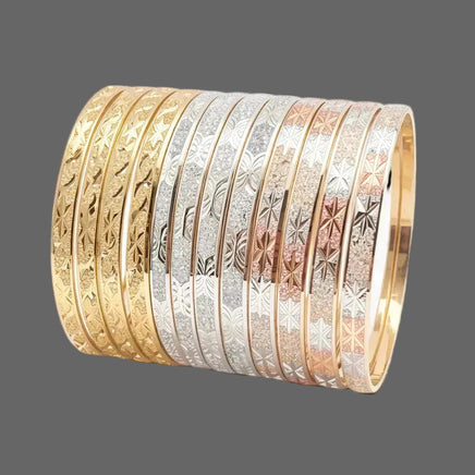 A stack of nine bangles in different metal finishes, including yellow gold, silver, rose gold, and white gold, each featuring an engraved floral pattern on the outer surface, displayed on a clean white background.